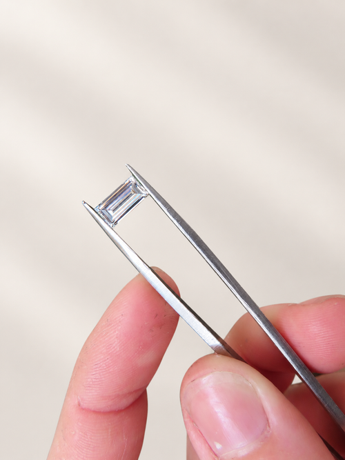 A close-up of a hand holding a silver tweezer with a rectangular clear gemstone between the metal tips, against a soft beige background.