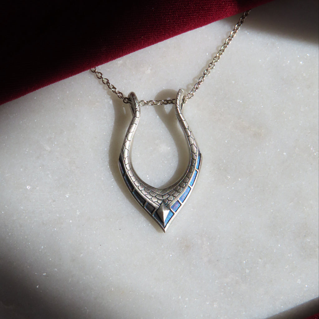 A silver horseshoe pendant necklace with blue detailing, resting on a white marble slab with a dark red fabric background.