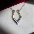 A silver horseshoe pendant necklace with blue detailing, resting on a white marble slab with a dark red fabric background.