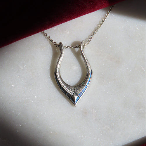 A silver horseshoe pendant necklace with blue detailing, resting on a white marble slab with a dark red fabric background.