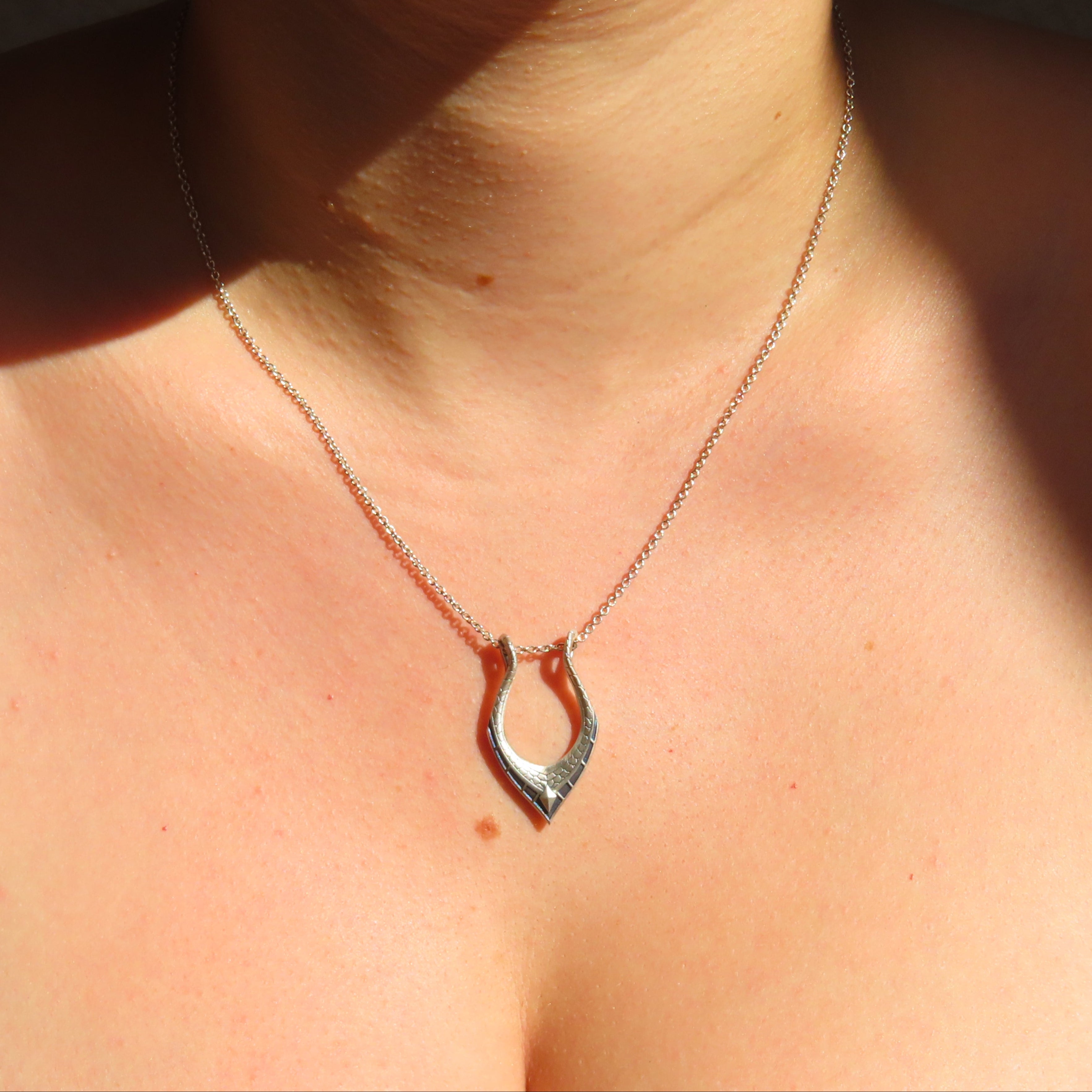 A close-up of a person's neck and chest wearing a silver necklace with a small, open, horsebit-like pendant.