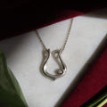 A silver-toned open lyre pendant necklace with a small clear gemstone, displayed on a white textured surface with dark red fabric and a green leaf in the background.
