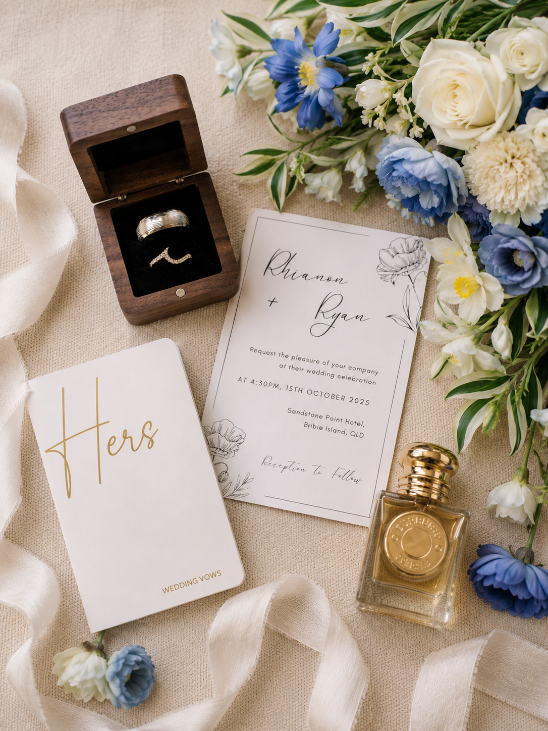 A styled flat-lay wedding scene on a beige fabric background with a wooden ring box containing two rings, a white wedding invitation card, a white 'Hers' card with gold script, a small perfume bottle, and blue and white flowers.