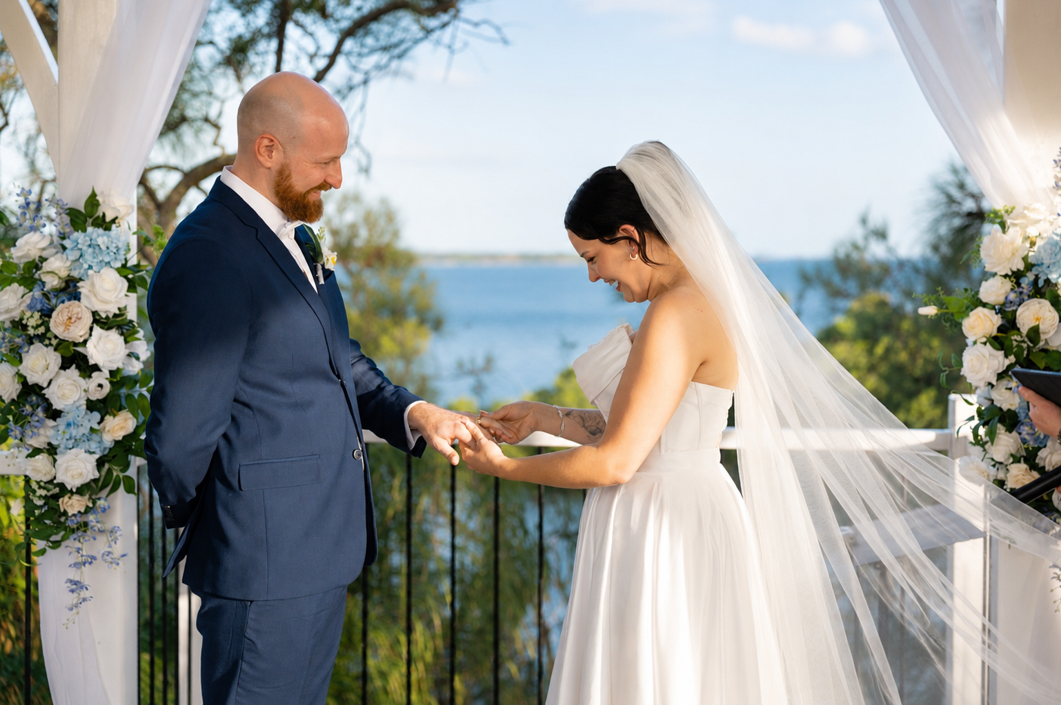A bride in a white wedding gown and veil and a groom in a navy suit exchanging rings under a white draped arch with floral arrangements, with a scenic ocean background.