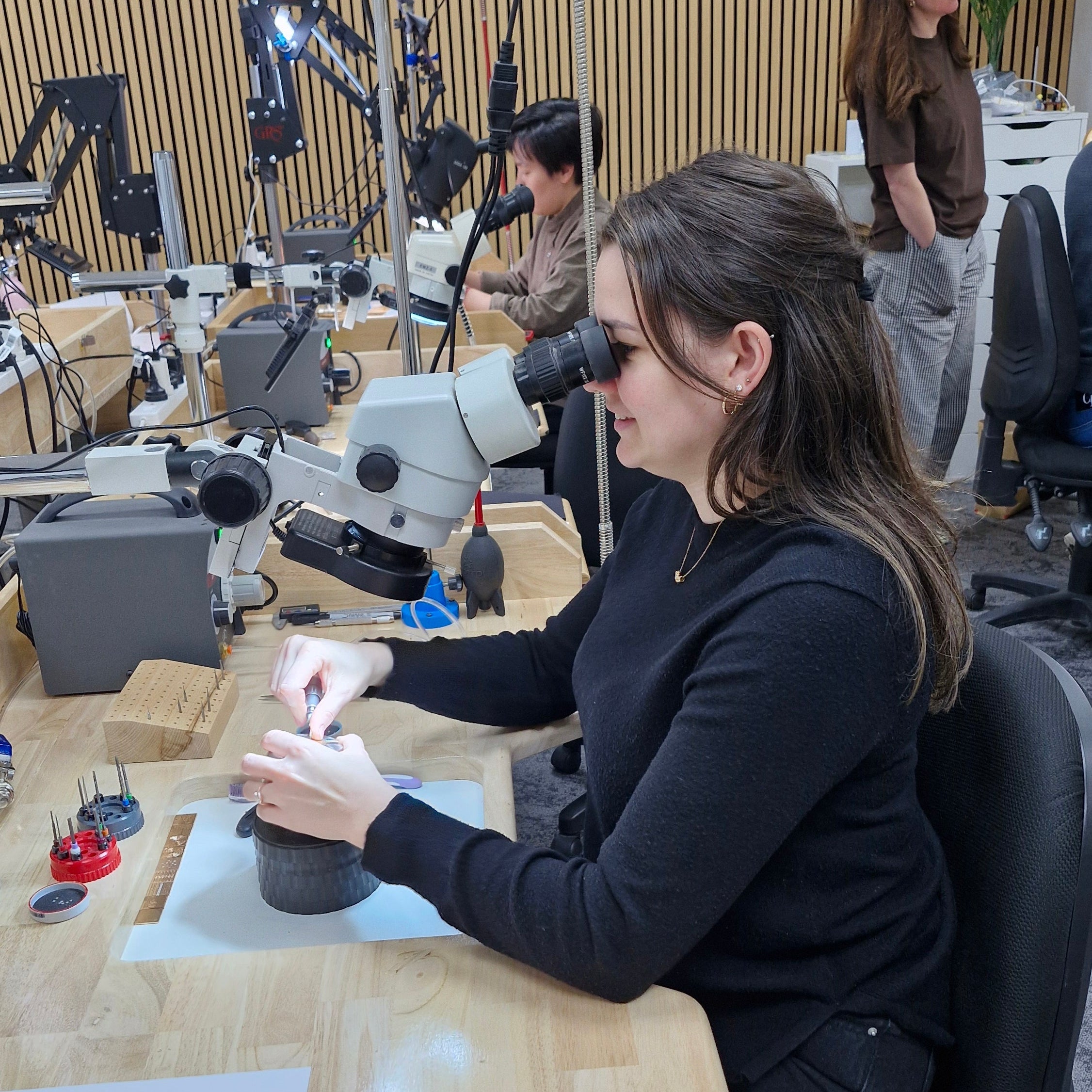 A woman in a black long-sleeve top works at a wooden desk under a microscope in a modern workshop or lab setting with other people and equipment in the background.