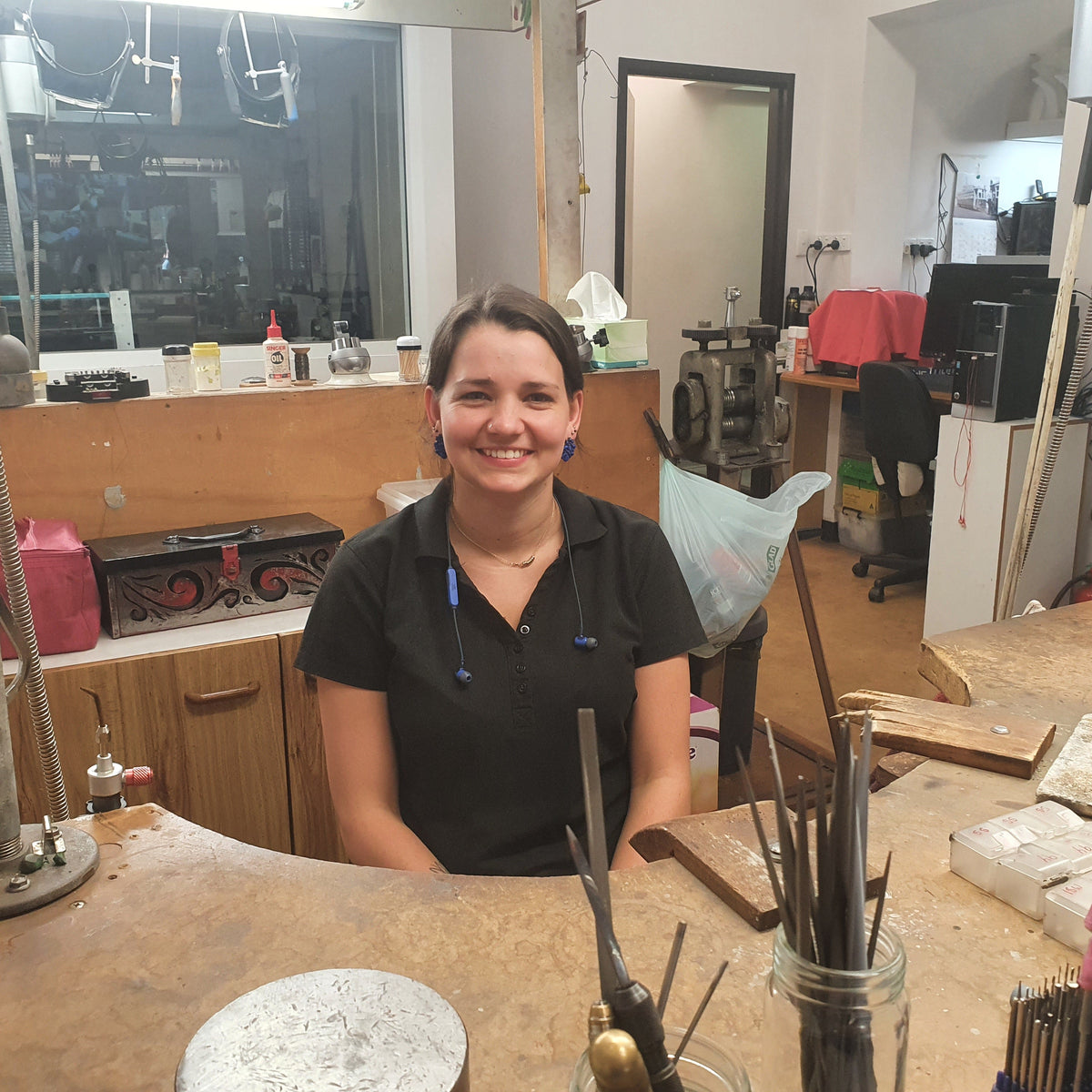 A smiling woman with dark hair tied back sits behind a wooden workbench in a workshop filled with tools, brushes, and equipment.