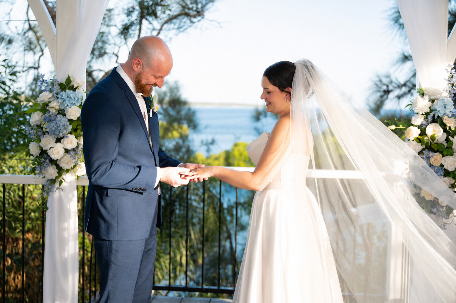 A bride in a white wedding gown and veil holding hands with a groom in a navy blue suit at an outdoor wedding under a white draped arch with floral arrangements.