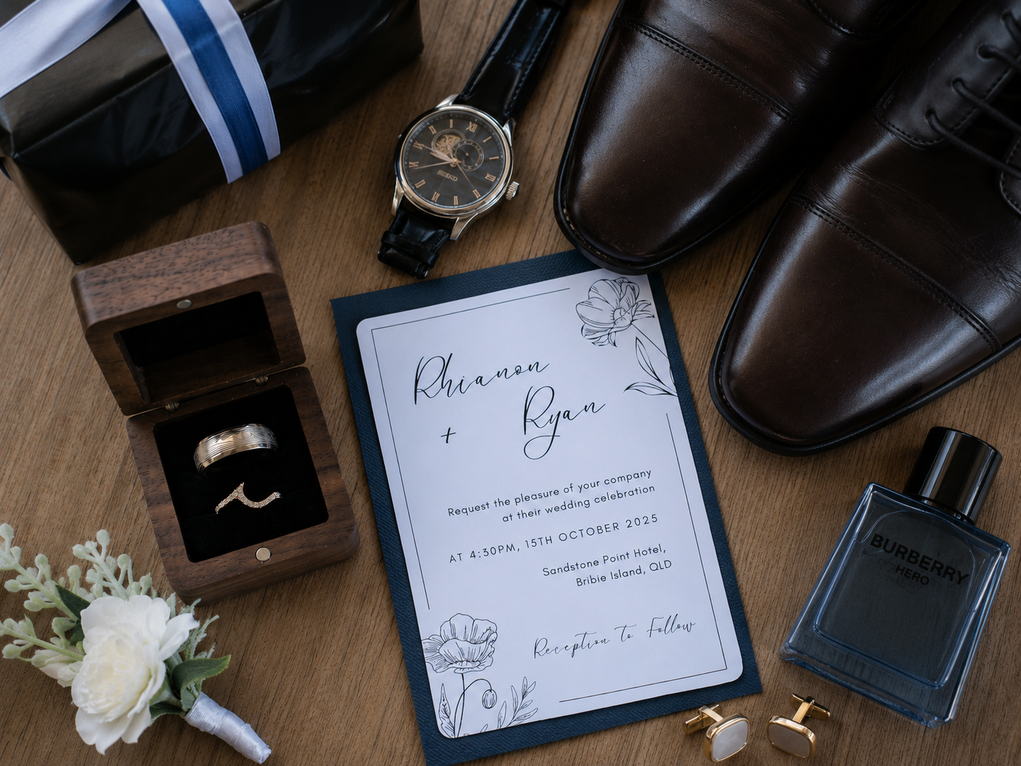 A styled flat-lay of wedding and formal accessories on a wooden surface, including a navy invitation card with floral script, a ring box with a gold band, a black dress watch, dark brown dress shoes, a Burberry bottle, cufflinks, and a white rose boutonniere.