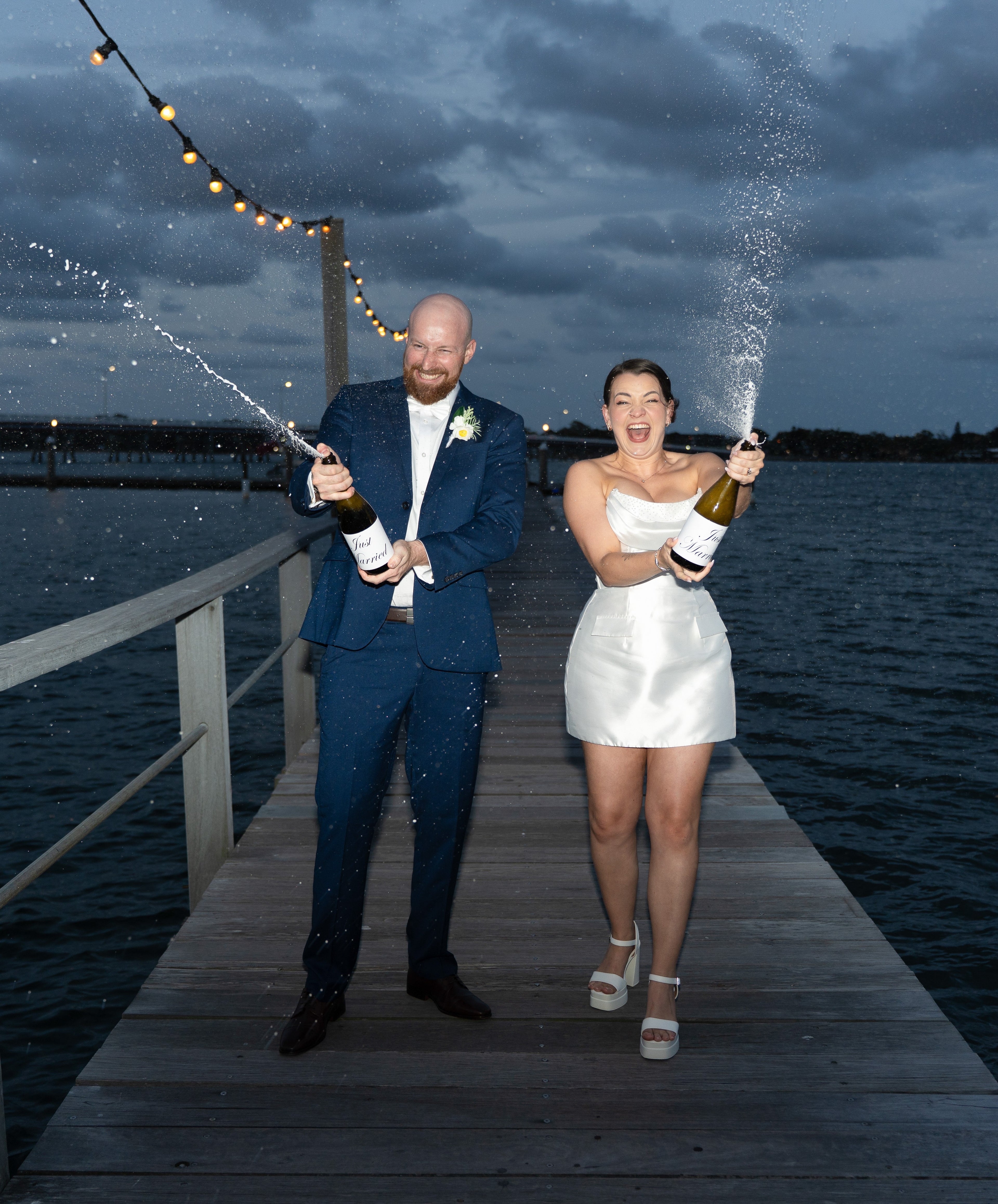 A bride and groom on a wooden dock over water, each holding a champagne bottle that is spraying foam. The groom is wearing a navy blue suit, and the bride is wearing a short white satin dress.