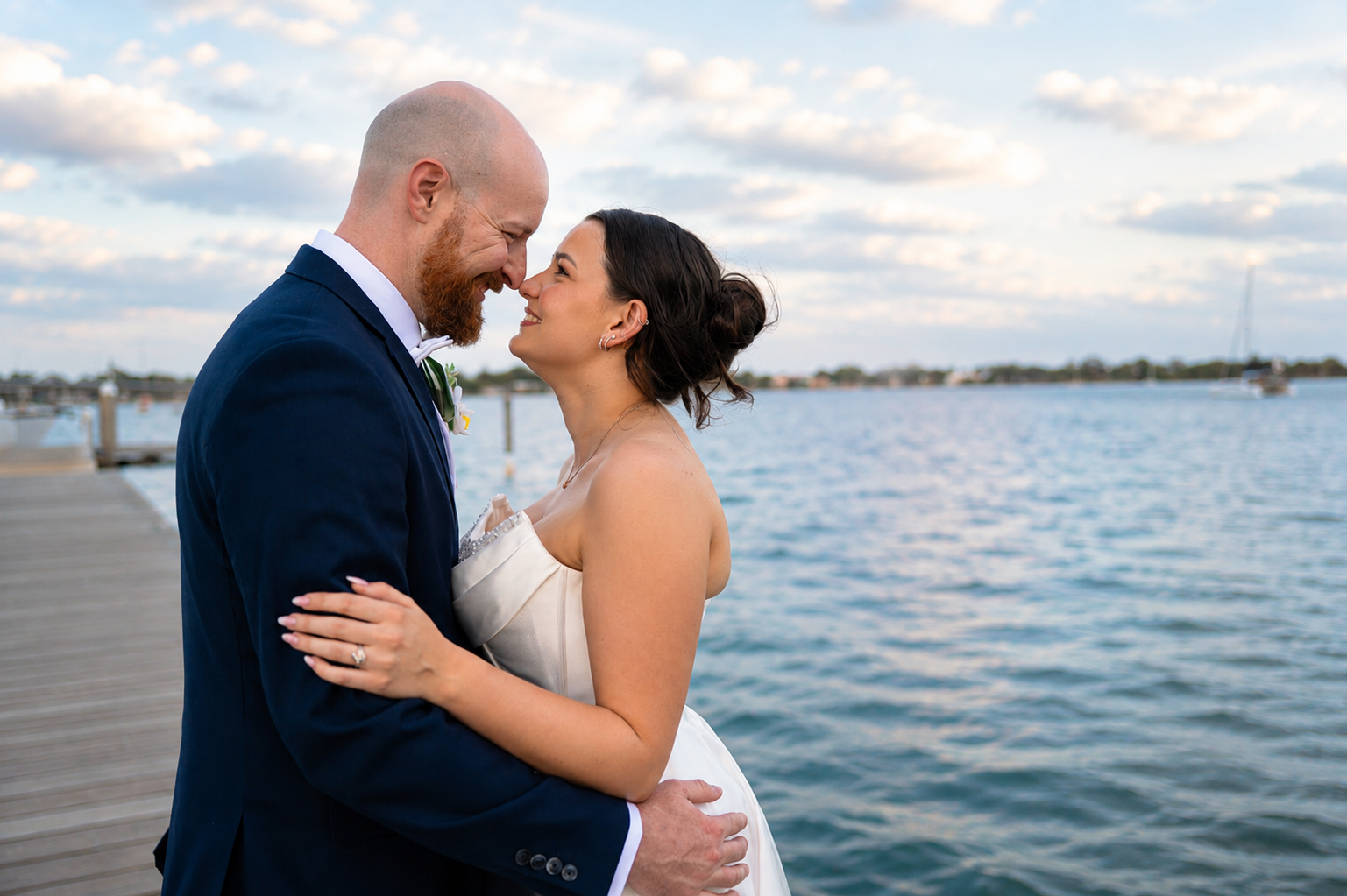 A bride in a white wedding gown and a groom in a navy blue suit embrace on a wooden dock with a scenic lake and cloudy sky in the background.
