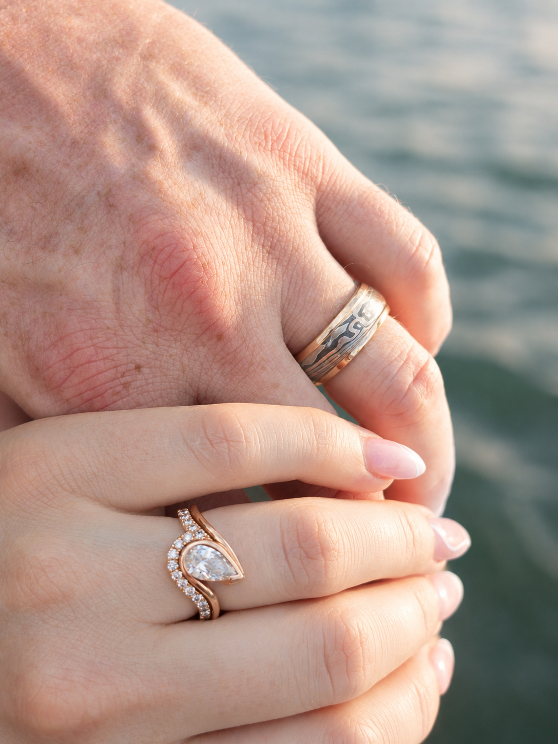 A close-up of two hands resting together outdoors, with the foreground hand wearing a rose gold ring featuring a pear-cut clear gemstone and small pavé accents, while the background hand wears a silver band ring.