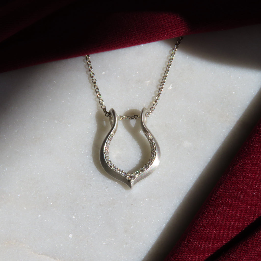 A silver horseshoe pendant necklace with a pavé-set clear stone border, resting on a white marble slab with dark red fabric draped in the background.